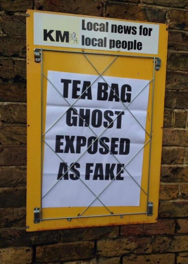 A local street advertisement displaying a provincial funny news headline from the KM news group pinned inside a wire-frame message box against a brick wall, declaring in massive block text: "TEA BAG GHOST EXPOSED AS FAKE."