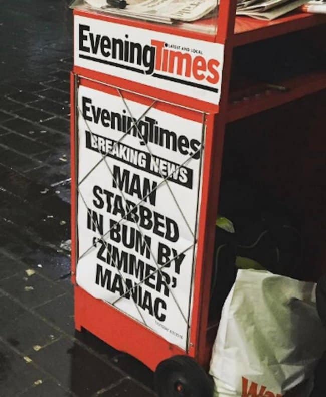 A roadside newspaper distribution cart displaying a highly alarming yet funny news headline poster from the Evening Times that reads "MAN STABBED IN BUM BY 'ZIMMER' MANIAC," drawing immense public attention to a hyper-specific and uncomfortable neighborhood assault incident.