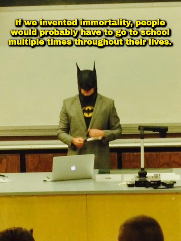funny shower thought meme displaying a man wearing a full Batman cowl paired with a grey business suit jacket over a Batman costume, standing behind a university lecture podium with a laptop, captioned: "If we invented immortality, people would probably have to go to school multiple times throughout their lives."