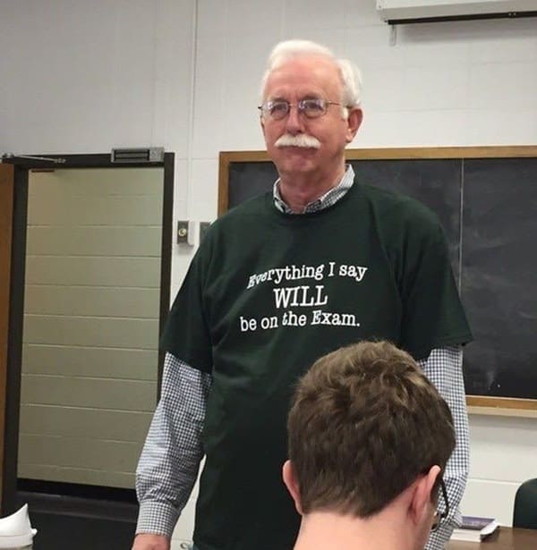 funny t shirt worn by an older male college professor with a thick white mustache standing in a classroom; the dark green shirt features bold white text stating, "Everything I say WILL be on the Exam."
