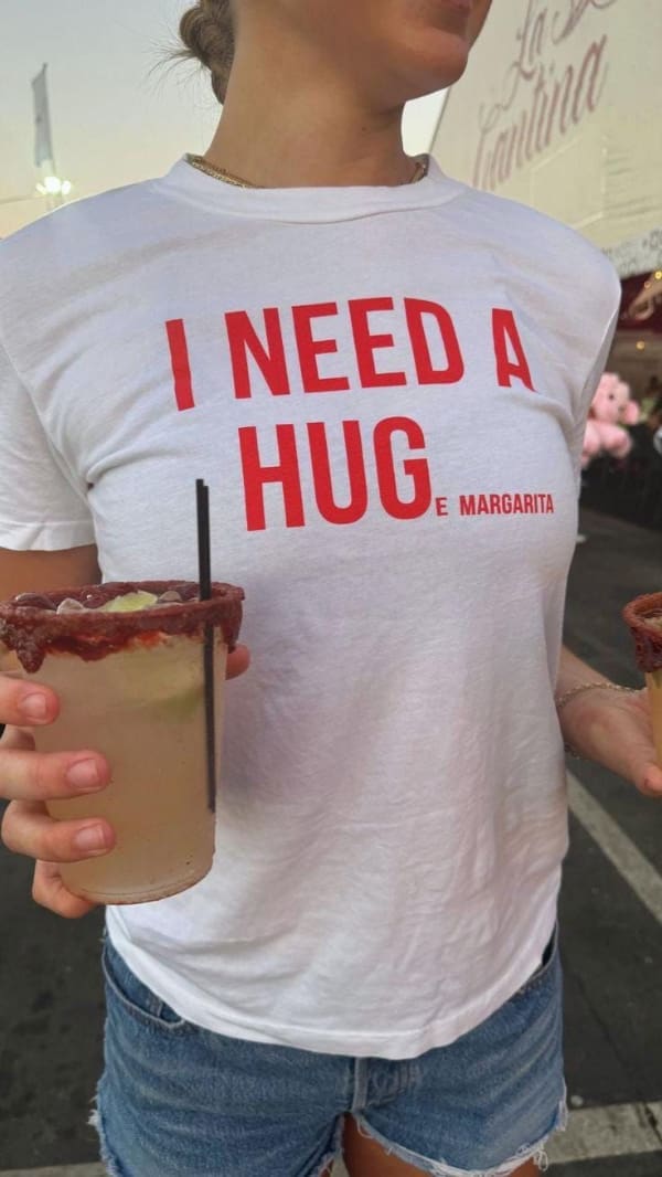 A clever funny t shirt worn by a woman holding a large rim-salted cocktail, featuring red lettering that initially reads "I NEED A HUG" but cleverly finishes in smaller print with "E MARGARITA" to read "I NEED A HUGE MARGARITA."