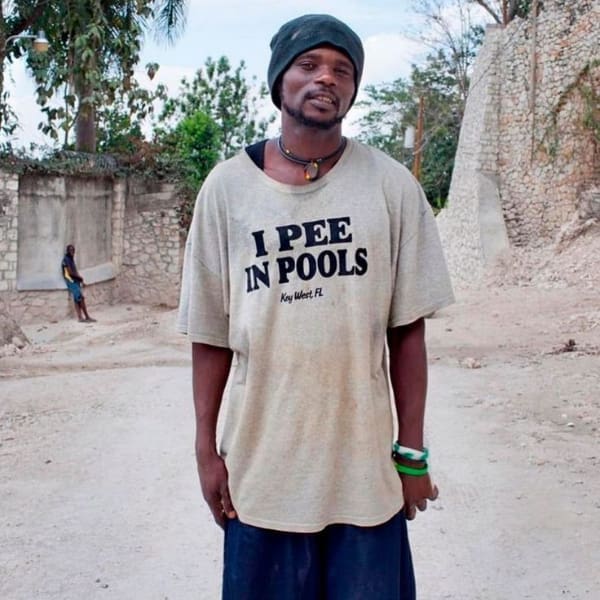 A funny t shirt worn by a man in a black beanie standing on a dirt road, proudly displaying bold black letters that confess his aquatic crimes: "I PEE IN POOLS Key West, FL."
