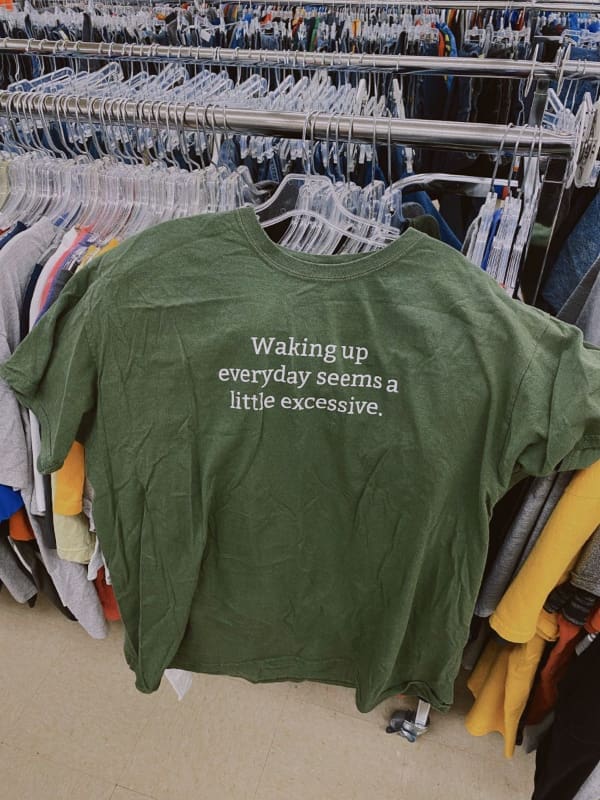 An incredibly relatable funny t shirt hanging on a thrift store rack, featuring a dark green fabric with crisp white text complaining: "Waking up everyday seems a little excessive."
