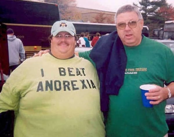 A classic funny t shirt modeled by a large, cheerful man standing outdoors next to a friend, utilizing bold stencil-style text on a light green background to loudly declare: "I BEAT ANOREXIA."