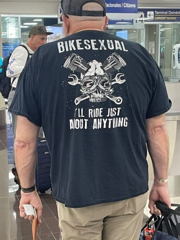 deeply unhinged biker funny t shirt captured from behind at an airport terminal, showcasing a large graphic of a skull and crossed wrenches under the loud, pun-heavy text: "BIKESEXUAL I'LL RIDE JUST ABOUT ANYTHING."