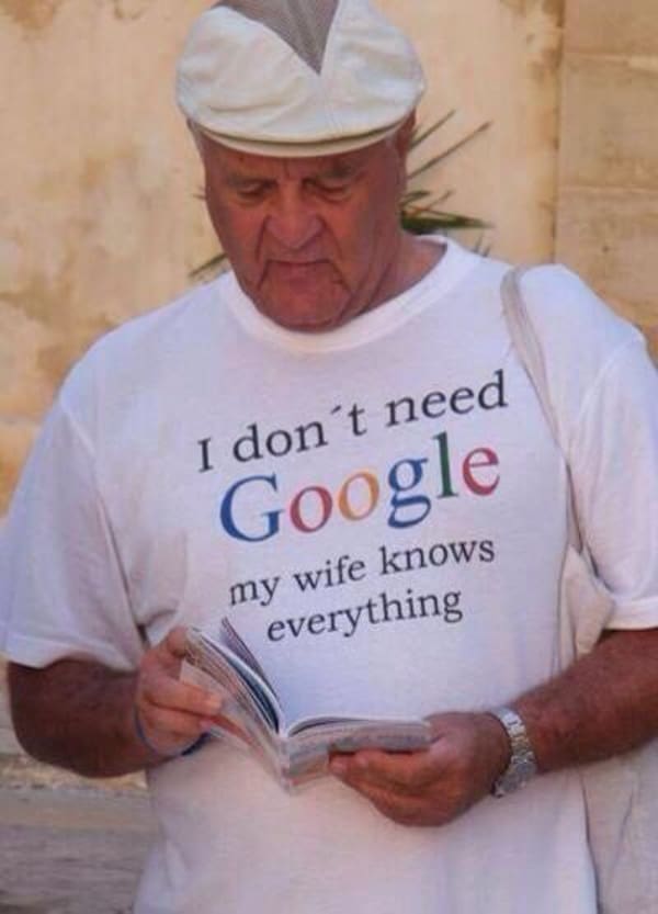 A boomer-humor funny t shirt modeled by an older man in a flat cap reading a guidebook, featuring the iconic colored search engine logo integrated seamlessly into the phrase: "I don't need Google my wife knows everything."