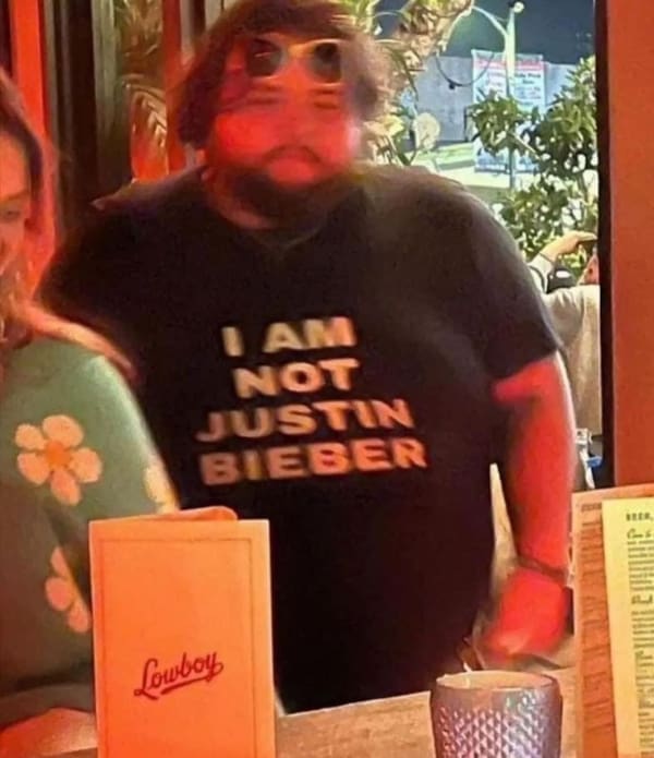 A deeply sarcastic funny t shirt worn by a large, heavily bearded man standing in a dimly lit bar, displaying the completely unnecessary but hilarious clarification: "I AM NOT JUSTIN BIEBER."