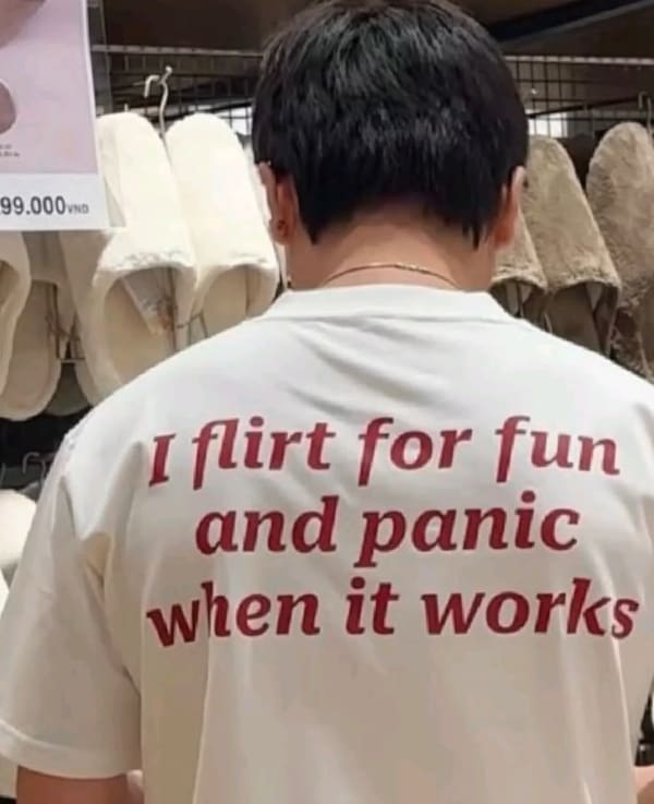 A relatable funny t shirt seen from behind on a person browsing a retail store, featuring sharp red serif text printed across the back that reads, "I flirt for fun and panic when it works."