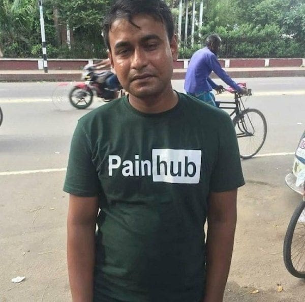 parody funny t shirt worn by a man standing outside on a city street, displaying a dark green shirt featuring a white-and-black logo that directly mimics a popular adult video website but reads "Painhub."
