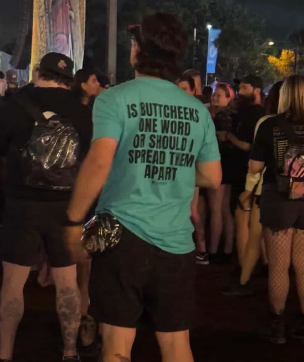 A crude yet funny t shirt captured from behind on a man standing in a crowded festival audience, featuring large black block lettering on a bright teal shirt that asks, "IS BUTTCHEEKS ONE WORD OR SHOULD I SPREAD THEM APART".