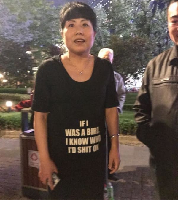 An aggressive funny t shirt worn by an older East Asian woman standing outside at night, displaying bold white capitalized lettering on a black shirt that reads, "IF I WAS A BIRD, I KNOW WHO I'D SH*T ON."