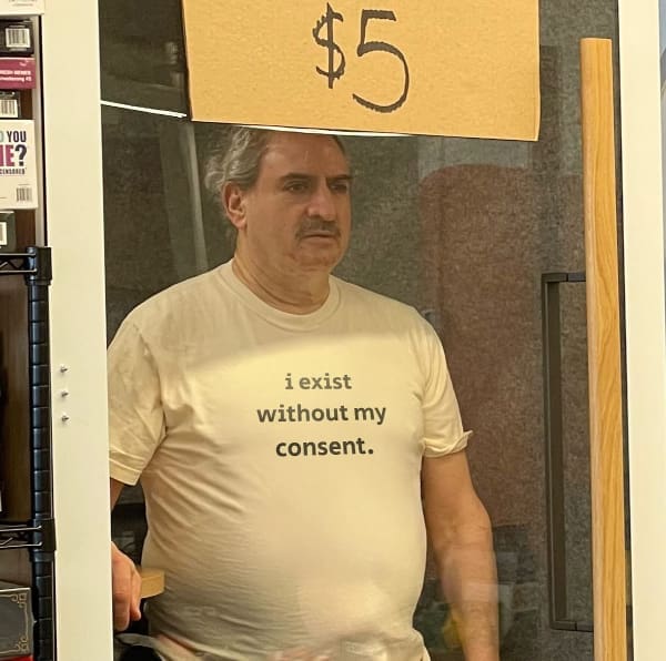 An existential funny t shirt worn by an older man with a mustache looking through a glass partition beneath a cardboard five-dollar sign; the shirt features simple lowercase text stating, "i exist without my consent."