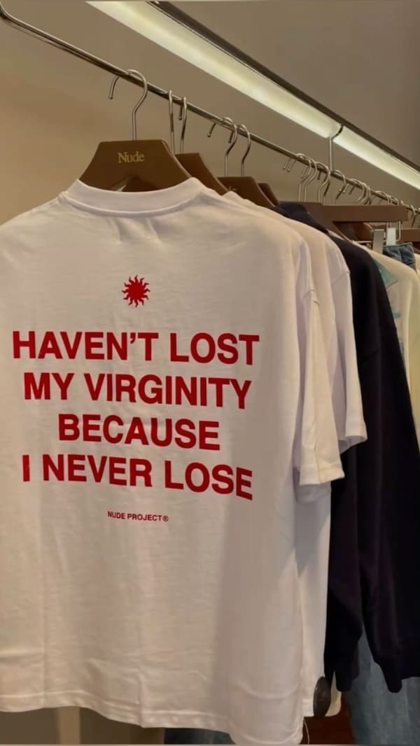 A competitive funny t shirt hanging on a clothing boutique rack from the brand Nude Project, featuring stark red block text that confidently proclaims, "HAVEN'T LOST MY VIRGINITY BECAUSE I NEVER LOSE."