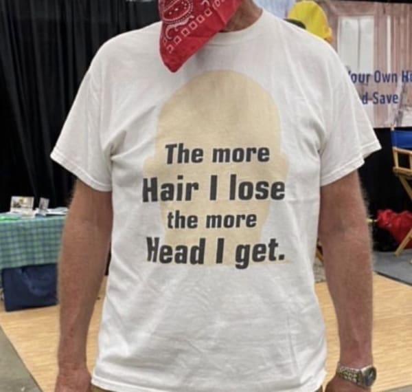 A bold funny t shirt worn by a man with a red bandana around his head, displaying a silhouette of a bald head alongside black text that reads, "The more Hair I lose the more Head I get."