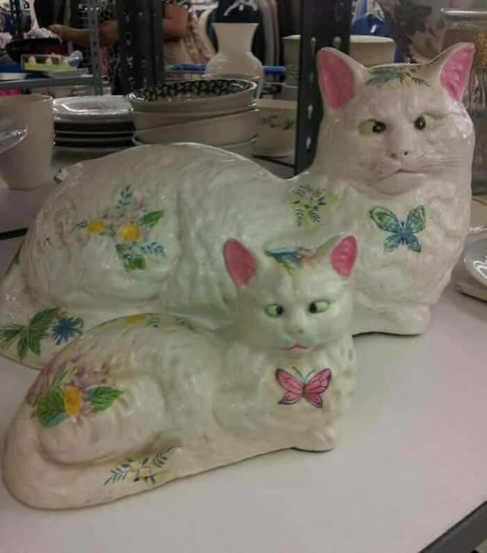 A poorly painted ceramic statue operating as a funny thrift store find, displaying a large mother cat and a smaller kitten adorned with pastel floral and butterfly decals, but their painted eyes are completely misaligned, giving both felines a hilariously deranged, cross-eyed expression.