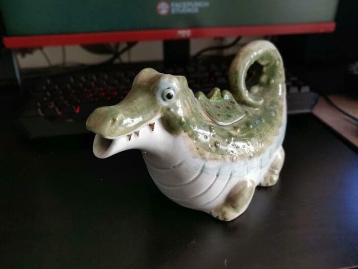 A peculiar piece of home decor serving as a funny thrift store find, showing a green ceramic pitcher specifically molded to look like a small alligator, where the creature's wide-open jaws function as the pouring spout.