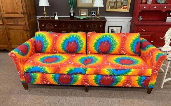 An incredibly loud piece of furniture acting as a funny thrift store find, displaying a full-sized living room sofa completely upholstered in a dizzying, ultra-vibrant, neon rainbow tie-dye fabric pattern.