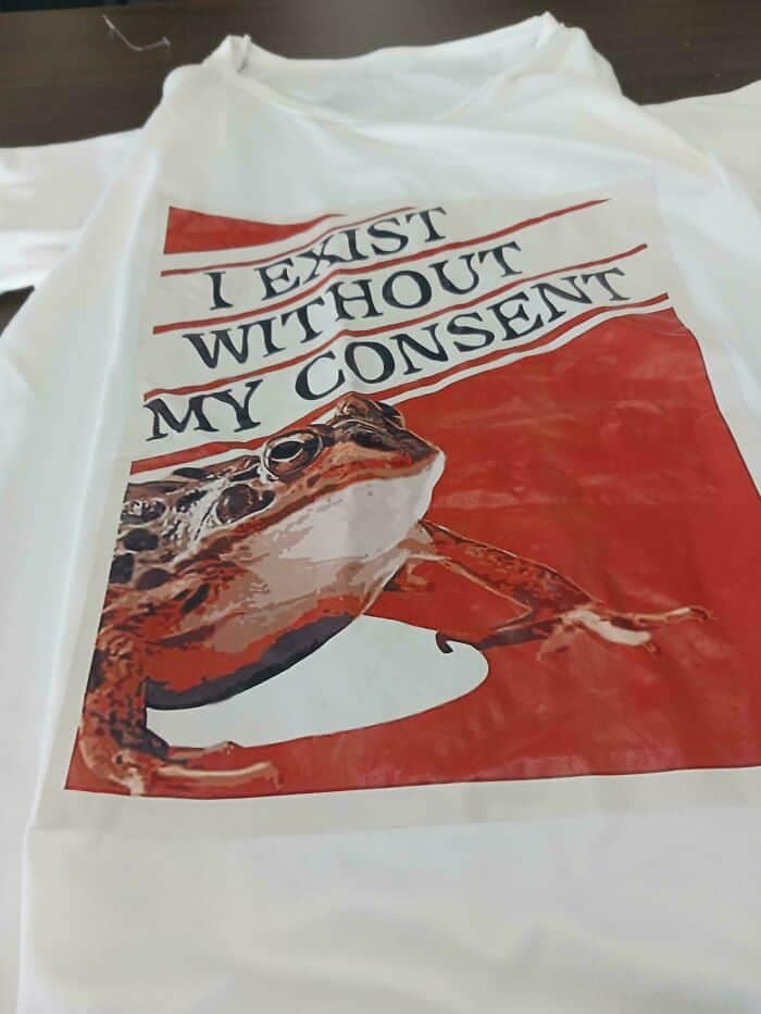 A chaotic graphic tee presented as a funny thrift store find, featuring a low-resolution image of a large green frog set against a bright red background, overlaid with the bold text: "I EXIST WITHOUT MY CONSENT".