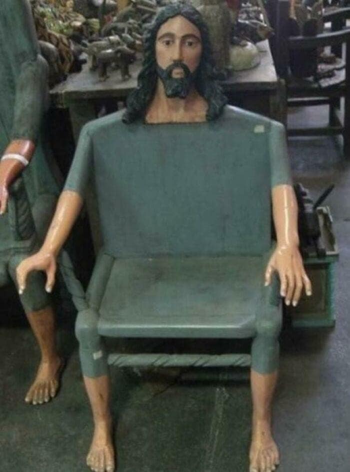 A highly unorthodox piece of religious furniture discovered as a funny thrift store find, showing a large wooden armchair painted dark green where the backrest extends upwards into a carved, realistic head of Jesus, while two wooden human arms rest on the armrests and two bare human feet serve as the front chair legs.
