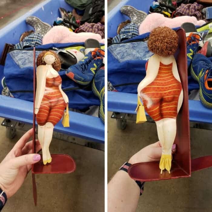 A highly specific funny thrift store find displaying a painted metal bookend shaped like a curvaceous woman with textured curly hair wearing a red striped dress, cleverly designed so that her prominent backside physically supports the books.