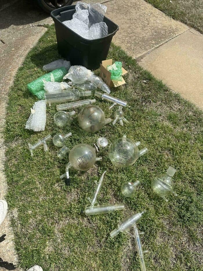 An extremely suspicious yard sale pile acting as a funny thrift store find, showing dozens of complex, intricate pieces of clear scientific chemistry glassware, round boiling flasks, and condenser tubes spread out on the grass next to a black storage bin.