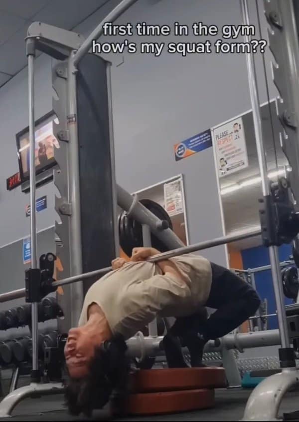 gym meme showing an absolute disaster of exercise execution where a beginner attempts a near-impossible back-bend bridge under a Smith machine barbell with their feet balanced on weight plates, captioned "first time in the gym how's my squat form??".
