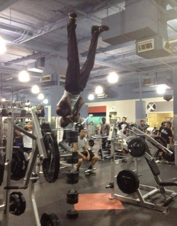 A gym meme capturing a jaw-dropping display of calisthenics balance where a fitness enthusiast performs a vertical, straight-legged handstand entirely balanced on top of a single tower of stacked dumbbells inside a commercial gym.