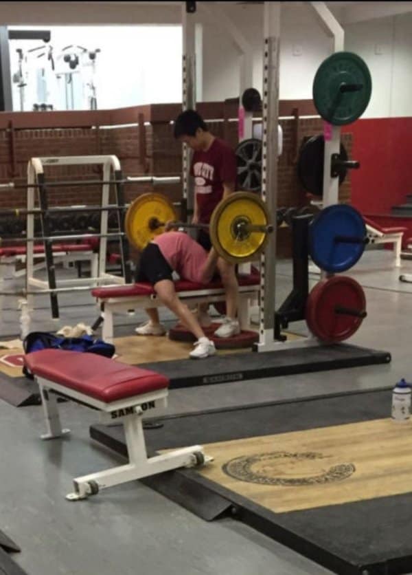 A gym meme capturing a powerlifter executing a heavy barbell bench press with an extreme, controversial spinal arch while a spotter looks on from above.