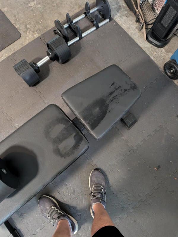 gym meme top-down photograph showcasing an adjustable home weight bench covered in a massive sweat stain that leaves a perfect, unmistakable silhouette of the lifter's backside.