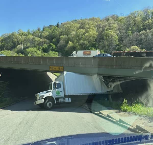 A white box truck wedged tightly underneath a low concrete highway overpass bridge.