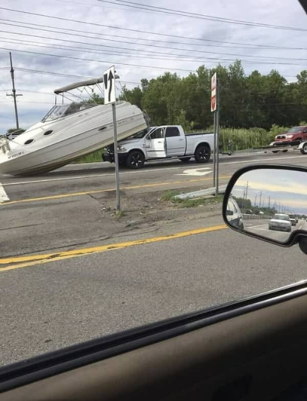 A large white pickup truck with a giant boat crashed onto its hood on a roadway.