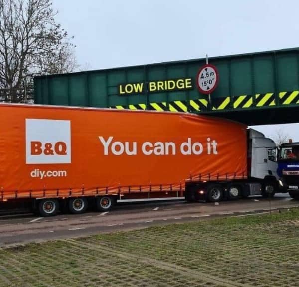 An orange semi-truck with a slogan reading you can do it stuck under a green low bridge.