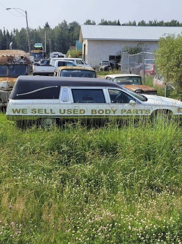 A vintage white hearse parked in a scrapyard with large text reading we sell used body parts.