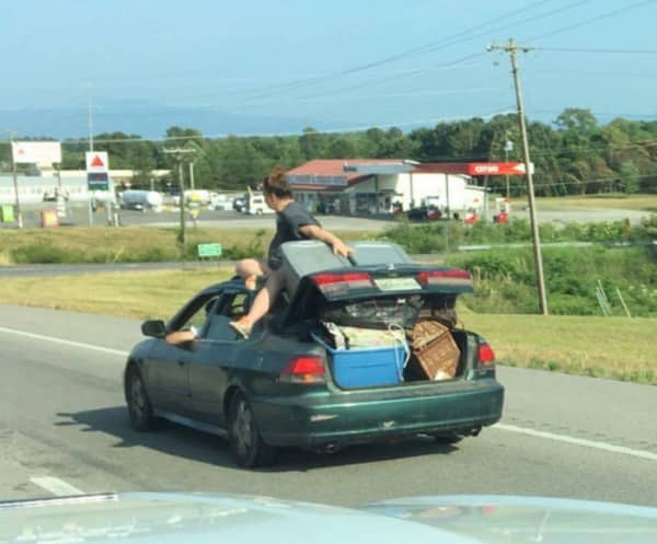 A woman sitting on the rear window of a car holding down an overflowing open trunk.
