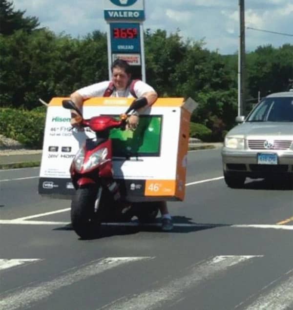 A man riding a small red moped while carrying a massive flat-screen television box on the handlebars.