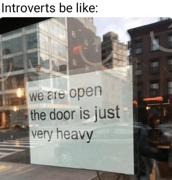 A real-world commercial storefront window sign reading "we are open the door is just very heavy," repurposed into a perfect visual metaphor for an introvert meme about personal accessibility boundaries.
