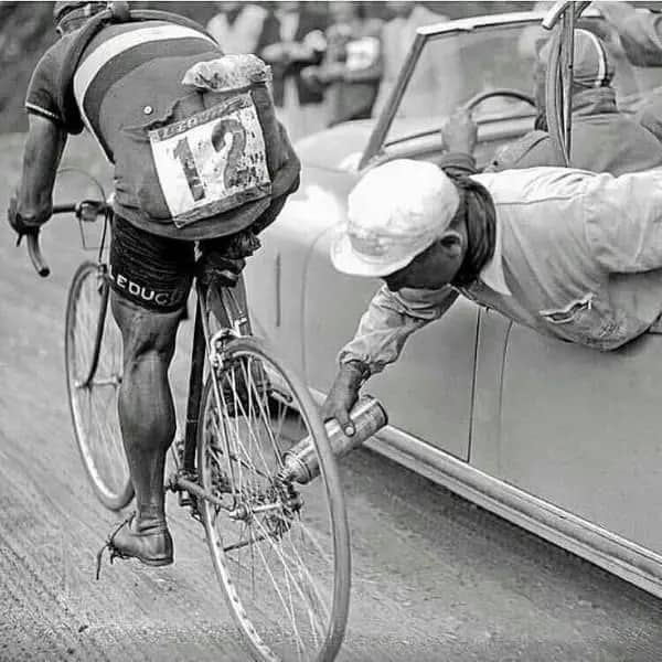 A vintage cyclist receiving a spray of liquid on their rear wheel from a moving car.
