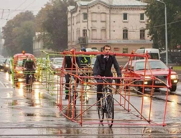 Cyclists riding on a wet street surrounded by large rectangular cage structures made of PVC pipes.