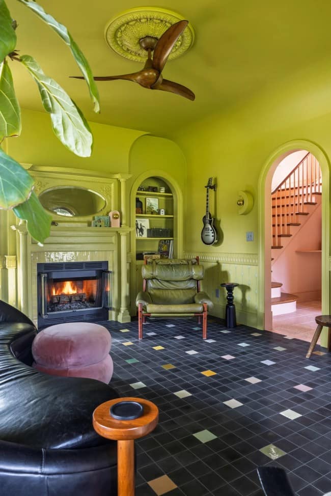 An example of striking maximalist interior design featuring a living room completely drenched in monochromatic chartreuse lime green from the walls and ceiling to the fireplace mantle. The space features a sculptural wooden ceiling fan, a vintage green leather armchair nestled into an arched alcove shelf, and a dark tiled floor accented with sporadic colored tiles.