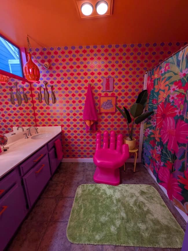 A vibrant, high-contrast bathroom executing loud maximalist interior design, featuring a shocking pink and orange trellis wallpaper, bright lavender vanity cabinets, a lime green shaggy rug, and a giant hot-pink hand-shaped plastic accent chair.