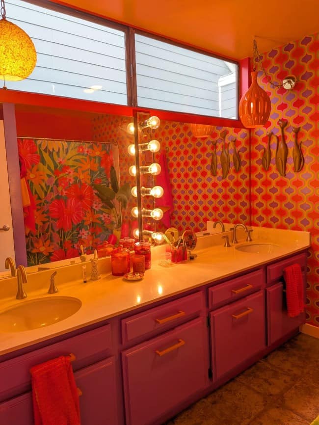 A double-sink bathroom vanity vanity unit executing bold maximalist interior design with rich magenta-painted cabinets, golden brass plumbing fixtures, dazzling Hollywood-style bulb strips, and a vibrant pink and orange geometric trellis wallpaper.