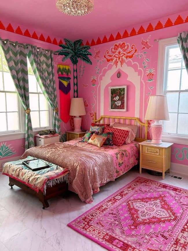 A whimsical tropical bedroom illustrating maximalist interior design with bubblegum pink walls, a painted green palm tree mural, graphic chevron-patterned green curtains, and a rich magenta traditional medallion area rug.