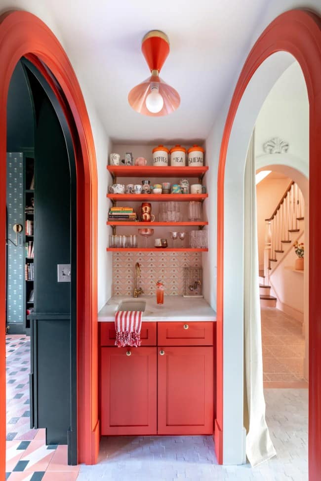 A recessed dry bar alcove demonstrating functional maximalist interior design with monochromatic tomato-red cabinets, matching painted arch trim, and floating wood shelves packed with an eclectic assortment of colorful mugs and coffee canisters.