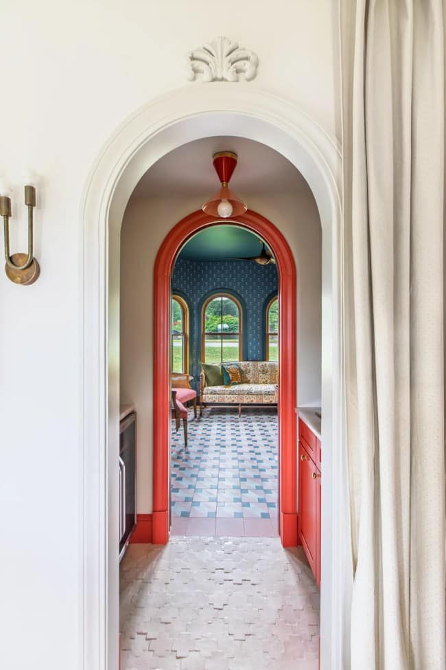 A layered architectural viewpoint tracing structural maximalist interior design, looking from a clean white hallway through a series of nested coral-red painted arches that frame a blue patterned sitting room with geometric tiled floors.
