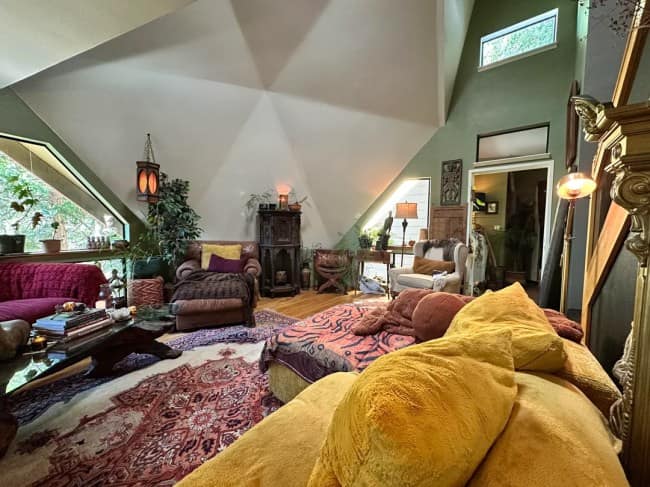 An attic loft apartment interior utilizing bohemian maximalist interior design inside a geodesic dome house, showcasing multi-angled white ceiling panels, a massive mustard-yellow velvet sectional sofa, and layered vintage Persian floor rugs.