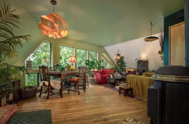 A spacious, open-concept dome house layout executing rustic maximalist interior design, featuring a rich hardwood floor, an expansive wooden dining table beneath a sculptural layered timber pendant light, a vintage wood stove, and a jungle of potted indoor plants.