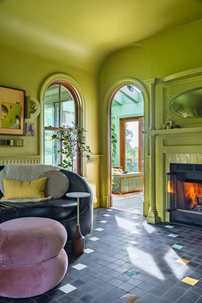 A continuation of the vibrant chartreuse living room showcasing maximalist interior design, detailing a plush pink velvet pouf resting on a black grid-patterned tile floor, a curved black leather sofa, and an open stone archway looking into a sun-soaked teal green sunroom.