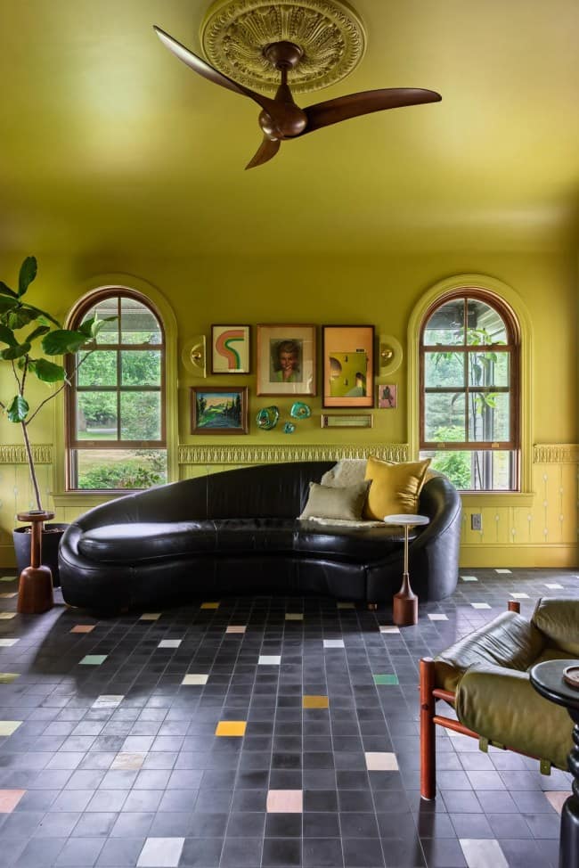 A symmetrical wide shot of a chartreuse green living room celebrating maximalist interior design, highlighting a large, curved black leather sofa centered between two elegant arched wooden windows under a gallery wall of eccentric abstract prints.