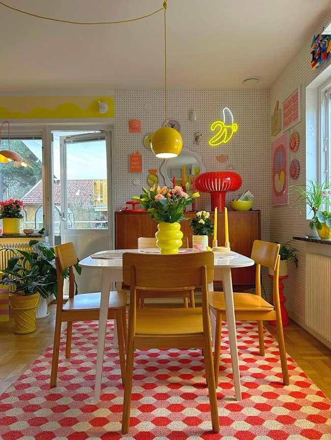 A whimsical, eclectic dining area exemplifying maximalist interior design with white polka-dot walls, a bright yellow pendant light matching a mod yellow table vase, a red plastic mushroom lamp on a teak sideboard, and a geometric red and white 3D-effect rug.