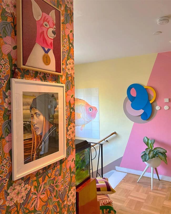 A colorful hallway landing exhibiting maximalist interior design with one wall covered in dense orange floral wallpaper holding a framed print of a chihuahua wearing a pink balaclava, alongside a yellow and pink color-blocked wall decorated with a blue abstract wood sculpture.
